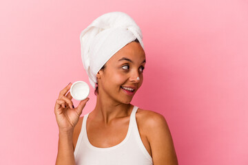 Young mixed race woman holding facial cream isolated on pink background looks aside smiling, cheerful and pleasant.