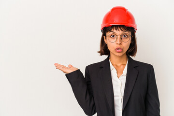 Young architect woman with red helmet isolated on white background impressed holding copy space on palm.