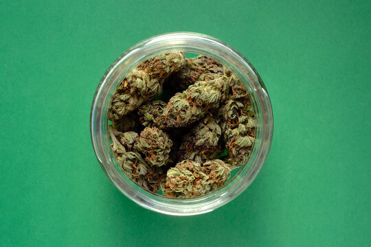 Close up of cbd cannabis buds in glass jar, macro view. Medicinal marijuana blooms on emerald green paper background. Hemp drying flowers, top view.