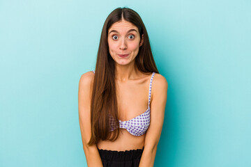 Young caucasian woman wearing bikini isolated on blue background shrugs shoulders and open eyes...