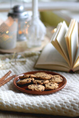 Plate of chocolate chip cookies, cinnamon sticks, soft blanket, open book and lit candles. Hygge at home. Selective focus.