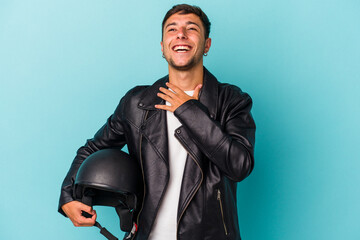 Young biker man holding helmet isolated on blue background  laughs out loudly keeping hand on chest.