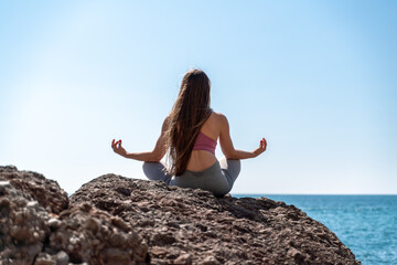 A young woman in gray leggings and a pink top with long loose hair does yoga outdoors by the sea on a sunny day. Women's yoga, fitness, Pilates. The concept of a healthy lifestyle, harmony.