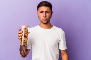Young caucasian man holding chocolate cookies isolated on purple background  shrugs shoulders and...
