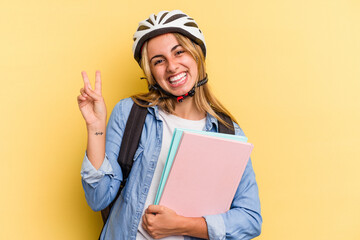 Young caucasian student woman wearing a bike helmet isolated on yellow background  joyful and carefree showing a peace symbol with fingers.
