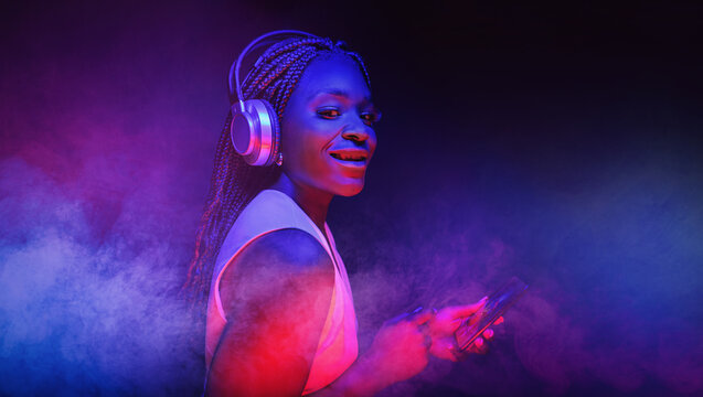  Young African American woman wearing headphones listening to music and dancing in futuristic purple cyberpunk neon light background