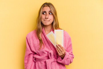 Young caucasian blonde woman wearing bathrobe holding depilatory bands isolated on yellow background  confused, feels doubtful and unsure.