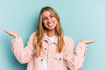Young caucasian blonde woman isolated on blue background  makes scale with arms, feels happy and confident.