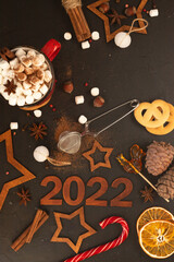 Wooden brown numbers 2022 on a dark background. There are stars carved out of wood, spices, scattered cocoa and a mug with marshmallows around