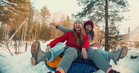 Two multiethnic excited happy beautiful friends women smile sledging on snow slope to camera,...