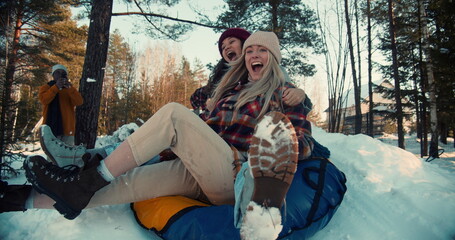Two attractive multiethnic happy women smile sledging on snow slope towards camera, winter weekend fun slow motion.