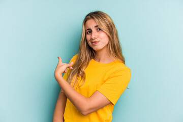 Young caucasian blonde woman isolated on blue background  smiling and pointing aside, showing something at blank space.