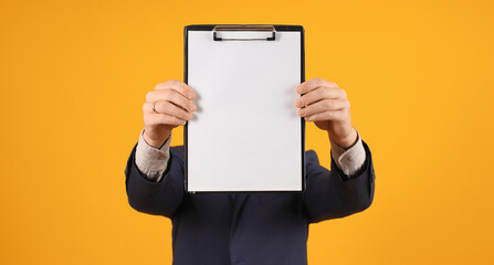 a man in a dark suit holds in front of him an empty tablet with a clip with a white sheet on a yellow background, mockup, close-up