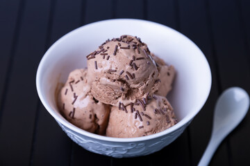 Chocolate ice cream with chocolate sprinkle in a white bowl on a black background. Three balls of homemade ice cream. Close-up.