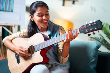 Slow motion shot of Happy Women enjoy singing by playing Guitar while sitting on sofa at home - Concept of Learning, practicing Guitar as hobby and Relaxing