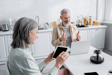 Mature man showing thumbs up during video call on laptop near coffee and wife with digital tablet at home.