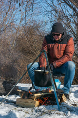 Caucasian man by campfire, pot of soot over bonfire hanging on tripod, winter outdoor cooking at campsite