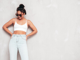 Young beautiful smiling female in trendy summer clothes. Sexy carefree woman posing near grey wall in studio. Positive brunette model having fun and going crazy. Cheerful and happy. In sunglasses