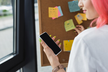 cropped view of businesswoman with pink hair holding smartphone with blank screen near board with sticky notes.