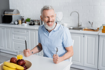 Smiling mature man holding cutlery near orange juice and fruits in kitchen.