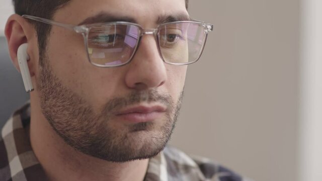 Slowmo close-up of serious bearded Mixed-race programmer in wireless earphones looking at computer screen while running program codes reflecting in his eyeglasses