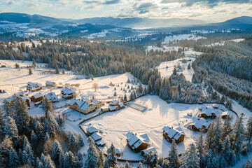 Aerial winter landscape with small village houses between snow covered forest in cold mountains in the evening