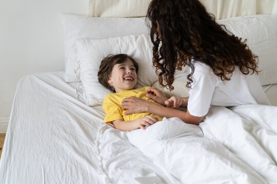 Cheering mother tickling cuddle little laughing son in bed after wake up on weekend morning. Loving mom and kid have fun in bedroom. Female parent bonding with preschool child at home. Love and family