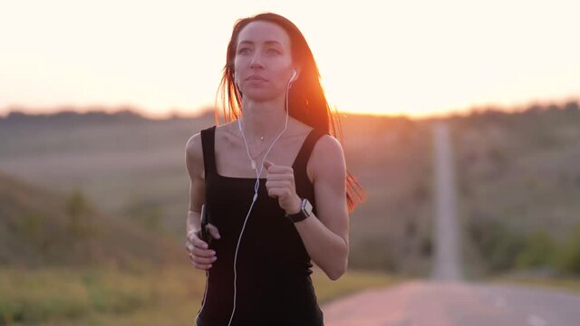 Concept healthy running. Young fitness woman running on road at sunset. Runner woman with fitness tracker and running app wearable technology with smart phone and earphones. Healthy lifestyle.