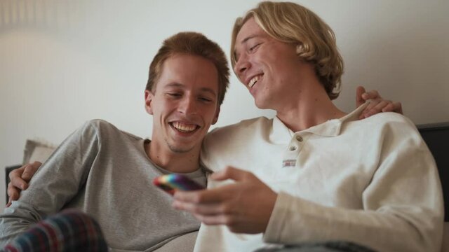 Laughing gay couple looking at the phone and talking while hugging on the bed