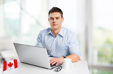 Happy young guy sitting with flag of Canada, using a laptop computer