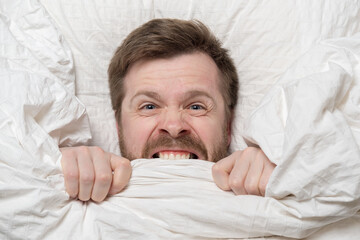 Caucasian man is under stress, suffers from insomnia, cannot sleep, nervously bites the blanket and...