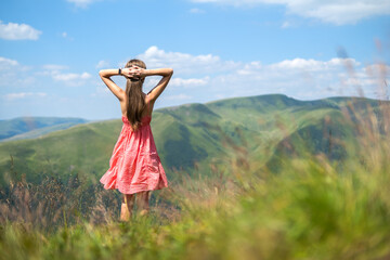 Young woman in red dress standing on grassy field on a windy day in summer mountains enjoying view of nature