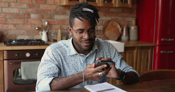 African Young Man In Glasses Sit At Table In Domestic Kitchen Holds Cellphone Share Voice Audio Message With Friend, Lead Conversation Using Speakerphone, Remote Communication, Modern App Tech Concept