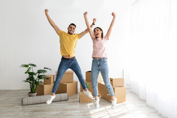 Excited multiracial couple celebrating relocation to their house, jumping among carton boxes on...