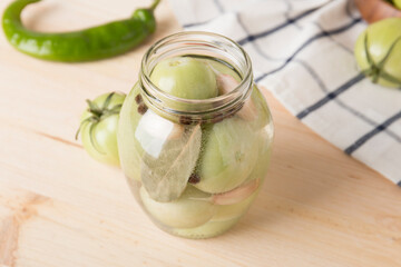 Green tomatoes for canning in a glass jar on a light wooden background. Unripe tomatoes for harvest.
