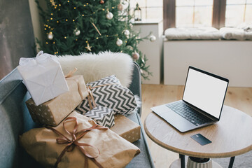 Laptop on table in interior with Christmas tree and presents. Holiday online shopping.
