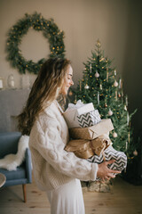 Young beautiful woman holding gift boxes and presents in a cozy room with Christmas tree on...