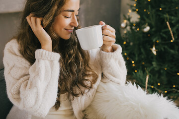 Young woman drinking hot coffee or cocoa sitting on couch in cozy room with Christmas tree. Winter...