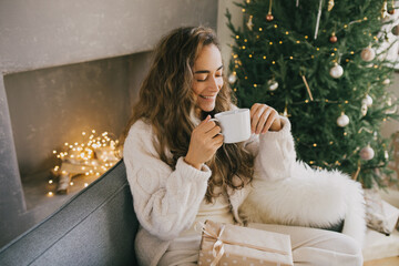 Young woman drinking hot coffee or cocoa sitting on couch in cozy room with Christmas tree. Winter holidays.
