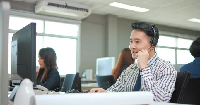 Asian man is customer support agent or call center with headset works on desktop computer while supporting the customer on phone call. 