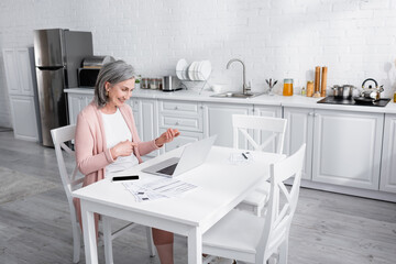 Cheerful mature woman pointing with fingers during video call on laptop near paper with bills in kitchen.