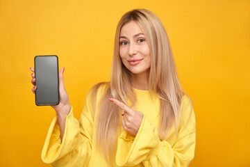 Naklejka premium Beautiful blonde girl pointing at blank phone screen and smiling isolated on yellow studio background
