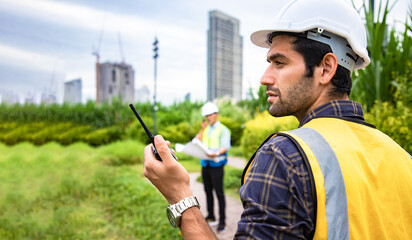 Portrait mature construction engineer confident with walkie talkie for check project and statistical report on site near cityscape. Back view of house property with laborer. Eco green