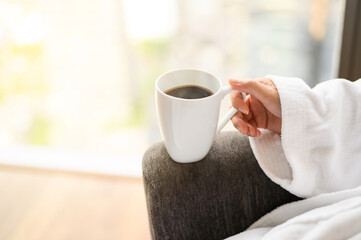 Side view of woman in bathrobe sitting near the window while holding white cup of coffee and enjoy wonderful morning in the hotel, detaching from work.