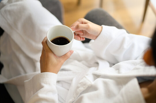 Top view of woman in bathrobe sitting near the window while holding white cup of coffee and enjoy wonderful morning in the hotel, detaching from work.