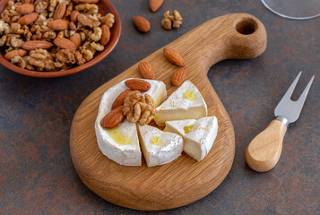 Small cheese platter. Sliced French brie or camembert cheese served on round wooden board with handle. Bowl walnuts and almonds on background. Selective focus.