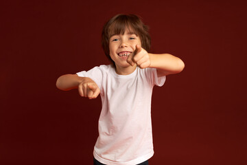 Cute, lovely, sweet adorable Caucasian kid with stylish haircut pointing at camera with both index fingers, fooling around, having fun, wearing white clothes standing against maroon background