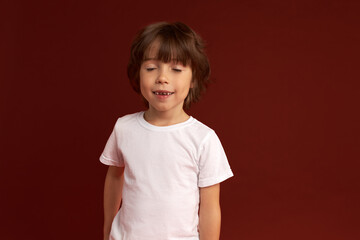 Close-up studio shot of handsome Caucasian brunette kid posing with eyes closed, smiling, dressed in white stylish t-shirt against maroon background with blank copy space for your text