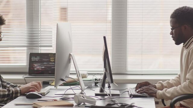 PAN shot of two multi-ethnic IT specialists sitting in front of each other in office writing program codes on computers