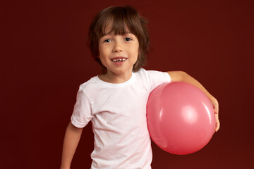 Studio image of charismatic photogenic kid of 7 years old carrying pink inflatable balloon in hand,...